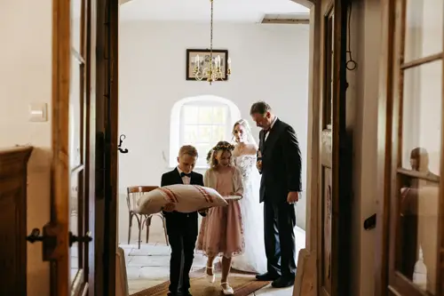 Ein kleiner Junge im Anzug und ein Mädchen in einem rosa Kleid mit Blumenkrone gehen gemeinsam mit einem großen Kissen den Gang einer Kirche entlang, während Hochzeitsgäste zuschauen und im Hintergrund die Braut mit ihrem Vater läuft.