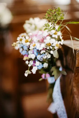 Ein kleiner, bunter Strauß Wildblumen, darunter weiße, rosa und blaue Blüten, ist mit einem Spitzenband an der Seite einer Holzbank befestigt, wahrscheinlich für eine Hochzeit oder einen besonderen Anlass. Der Hintergrund ist leicht verschwommen.