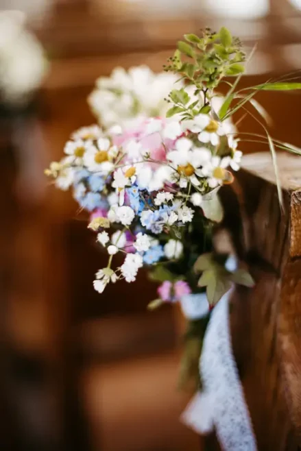 Ein kleiner, bunter Strauß Wildblumen, darunter weiße, rosa und blaue Blüten, ist mit einem Spitzenband an der Seite einer Holzbank befestigt, wahrscheinlich für eine Hochzeit oder einen besonderen Anlass. Der Hintergrund ist leicht verschwommen.