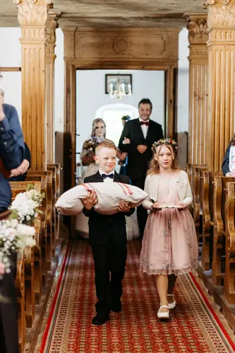 Ein kleiner Junge im Anzug und ein Mädchen in einem rosa Kleid mit Blumenkrone gehen gemeinsam mit einem großen Kissen den Gang einer Kirche entlang, während Hochzeitsgäste zuschauen und im Hintergrund die Braut mit ihrem Vater läuft.