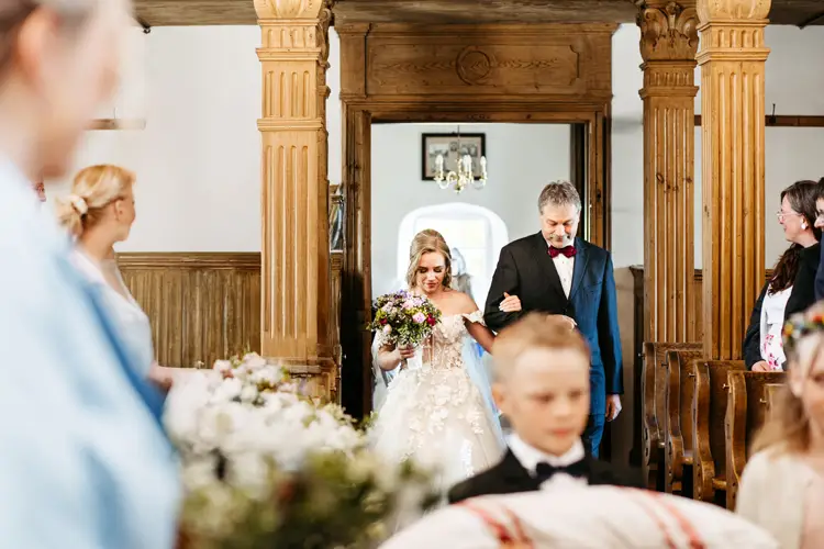 Eine Braut in einem weißen Kleid und einem Blumenstrauß in der Hand geht Arm in Arm mit einem älteren Mann im Anzug den Gang entlang, während die Gäste von den Holzbänken einer hellen Kirche aus zuschauen.