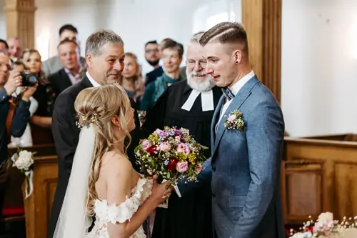 Braut und Bräutigam stehen sich in einer Kirche gegenüber, halten Händchen und lächeln. Die Braut hält einen Blumenstrauß in der Hand. Im Hintergrund schauen Gäste und ein Geistlicher in schwarzer Robe zu.