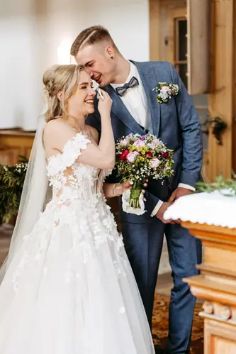 Eine lachende Braut in einem weißen Brautkleid mit Blumenmuster steht neben einem lächelnden Bräutigam in blauem Anzug und Fliege und hält einen bunten Blumenstrauß in einem warm beleuchteten Raum.