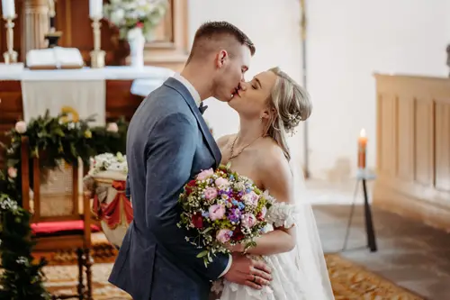 Braut und Bräutigam küssen sich in einer geschmückten Kirche. Die Braut hält einen bunten Blumenstrauß in der Hand und trägt ein weißes Kleid, der Bräutigam einen blauen Anzug. Im Hintergrund sind ein Altar und eine brennende Kerze zu sehen.
