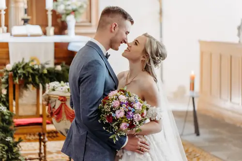 Braut und Bräutigam lächeln sich in einer geschmückten Kirche liebevoll an. Die Braut hält einen bunten Blumenstrauß in der Hand und trägt ein weißes Kleid, der Bräutigam einen blauen Anzug. Der Raum ist von sanftem Tageslicht erfüllt.