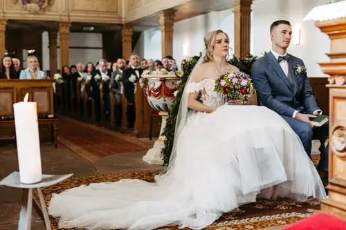 Eine Braut im weißen Kleid und ein Bräutigam im blauen Anzug sitzen nebeneinander in einer geschmückten Kirche und blicken beide nach vorne; die Braut hält einen bunten Blumenstrauß.