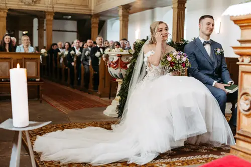 Eine Braut im weißen Kleid und ein Bräutigam im blauen Anzug sitzen nebeneinander in einer geschmückten Kirche und blicken beide nach vorne; die Braut hält einen bunten Blumenstrauß.