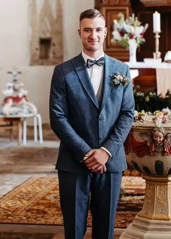 Ein lächelnder Mann in einem blauen Anzug mit Fliege und Blumenanstecker steht im Innenbereich, möglicherweise bei einer Hochzeitszeremonie, mit verschwommenem Kirchendekor im Hintergrund.