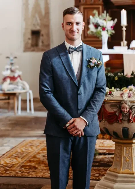 Ein lächelnder Mann in einem blauen Anzug mit Fliege und Blumenanstecker steht im Innenbereich, möglicherweise bei einer Hochzeitszeremonie, mit verschwommenem Kirchendekor im Hintergrund.