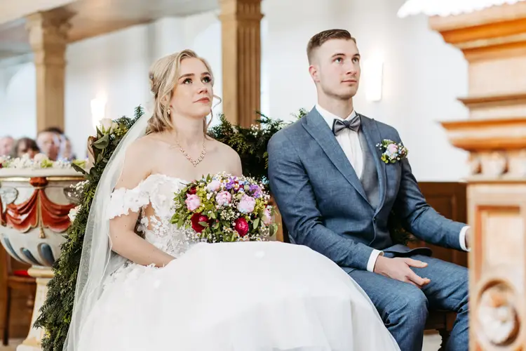 Eine Braut im weißen Kleid und ein Bräutigam im blauen Anzug sitzen nebeneinander in einer geschmückten Kirche und blicken beide nach vorne; die Braut hält einen bunten Blumenstrauß.