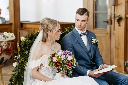 Eine Braut in weißem Kleid und Schleier sitzt neben ihrem Bräutigam im blauen Anzug in einer Kirche. Sie halten gemeinsam ein Buch in den Händen. Hinter ihnen sitzen Gäste in den Kirchenbänken. Im Hintergrund sind Blumendekorationen zu sehen.