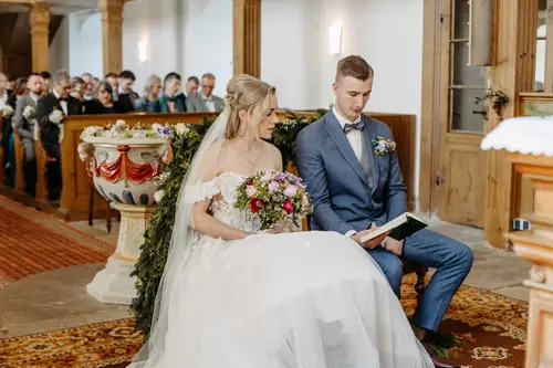 Eine Braut in weißem Kleid und Schleier sitzt neben ihrem Bräutigam im blauen Anzug in einer Kirche. Sie halten gemeinsam ein Buch in den Händen. Hinter ihnen sitzen Gäste in den Kirchenbänken. Im Hintergrund sind Blumendekorationen zu sehen.