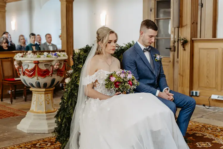 Eine Braut in weißem Kleid und Schleier sitzt neben einem Bräutigam im blauen Anzug während einer Hochzeitszeremonie in einer Kirche. Beide blicken ernst. Im Hintergrund sitzen Gäste, und Blumenschmuck ist zu sehen.