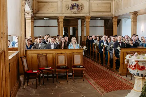 Eine Gruppe gut gekleideter Gäste sitzt in Holzbänken in einer Kirche mit kunstvoller Holzvertäfelung und nimmt an einer offiziellen Zeremonie teil. Einige halten Blumen in den Händen, alle blicken nach vorne und beobachten aufmerksam.