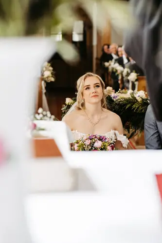 Eine Braut in einem weißen Kleid und Schleier hält einen Blumenstrauß und blickt nachdenklich nach oben, während sie bei einer Hochzeitszeremonie sitzt, mit verschwommenen Gästen und Blumendekorationen im Hintergrund.