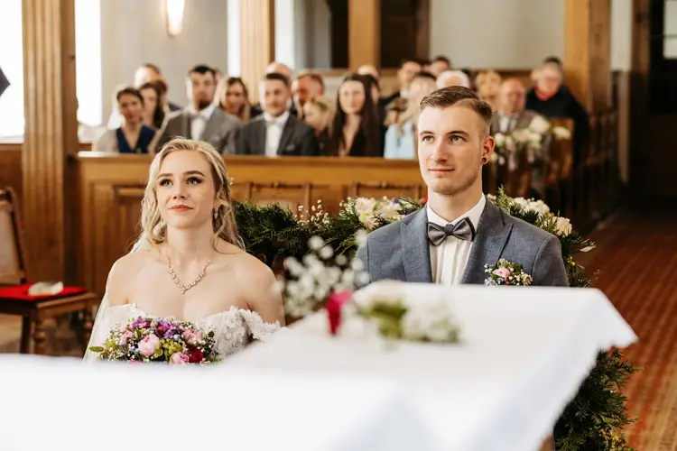 Während einer Hochzeitszeremonie sitzen Braut und Bräutigam Seite an Seite am Altar und halten bunte Blumensträuße in den Händen, während die Gäste in den Kirchenbänken sitzen und von hinten zuschauen.