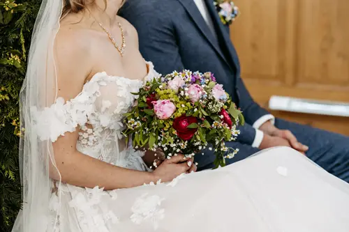 Eine Braut in einem weißen schulterfreien Brautkleid und Schleier hält einen bunten Blumenstrauß in der Hand und sitzt neben dem Bräutigam, der einen blauen Anzug trägt. Ihre Gesichter sind nicht zu sehen.