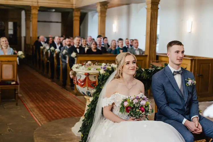 Braut und Bräutigam sitzen lächelnd und mit Blumensträußen in der Hand vor einer Kirche, während die Gäste in den Holzbänken der Hochzeitszeremonie zuschauen. Die Kirche ist mit Grünpflanzen und Blumen geschmückt.