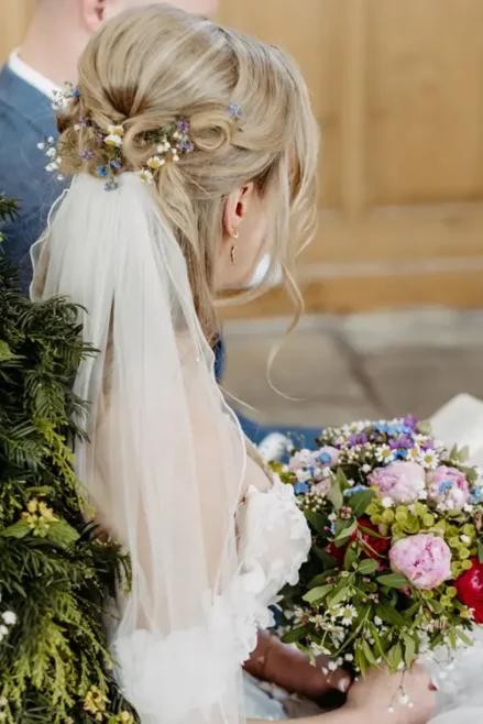 Eine Braut mit welligem blondem Haar, das zu einer Hochsteckfrisur mit kleinen Blumen und einem Schleier gestylt ist, hält einen bunten Strauß rosa, violetter und weißer Blumen in der Hand und sitzt neben Grünpflanzen.