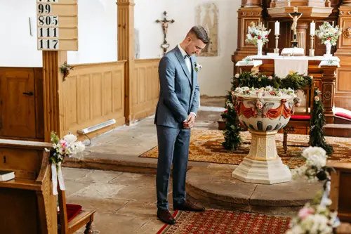 Ein Mann im blauen Anzug steht allein in einer geschmückten Kirche, mit gesenktem Kopf und gefalteten Händen, neben einem mit Blumen geschmückten Taufbecken. Im Hintergrund sind Holzbänke, Blumenarrangements und religiöse Symbole zu sehen.