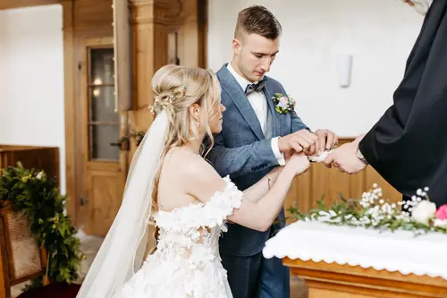 Eine Braut in einem weißen Kleid und ein Bräutigam in einem blauen Anzug tauschen während einer Hochzeitszeremonie in einer Kirche Ringe aus. In der Nähe befindet sich ein Priester, der die Zeremonie leitet, und Blumenschmuck ist angebracht.