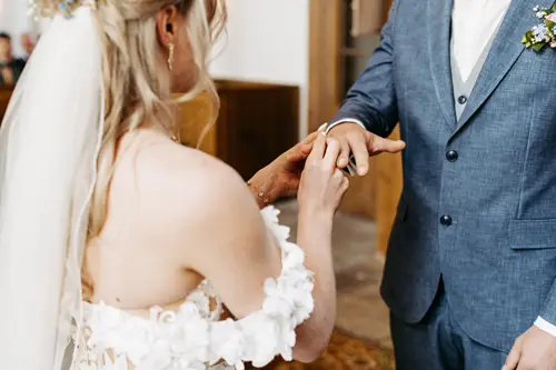 Braut und Bräutigam stehen sich im Innenbereich gegenüber, während die Braut dem Bräutigam sanft einen Ring an den Finger steckt. Die Braut trägt ein schulterfreies weißes Kleid und einen Schleier; der Bräutigam trägt einen blauen Anzug mit Fliege und Ansteckblume.