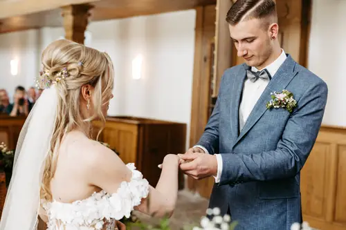 Braut und Bräutigam stehen sich im Innenbereich gegenüber, während der Bräutigam der Braut sanft einen Ring an den Finger steckt. Die Braut trägt ein schulterfreies weißes Kleid und einen Schleier; der Bräutigam trägt einen blauen Anzug mit Fliege und Ansteckblume.
