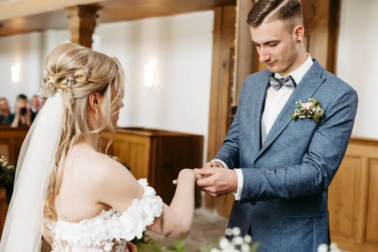 Braut und Bräutigam stehen sich im Innenbereich gegenüber, während der Bräutigam der Braut sanft einen Ring an den Finger steckt. Die Braut trägt ein schulterfreies weißes Kleid und einen Schleier; der Bräutigam trägt einen blauen Anzug mit Fliege und Ansteckblume.