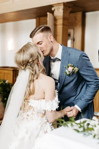 Braut und Bräutigam küssen sich drinnen. Die Braut trägt ein weißes, schulterfreies Kleid mit geblümtem Schleier, der Bräutigam einen blauen Anzug mit einer rosa Ansteckblume. Sie halten Händchen und lächeln.