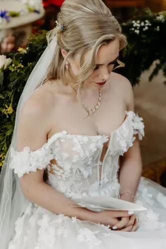Eine Braut mit blondem, welligem Haar und Schleier sowie einem schulterfreien, weißen Brautkleid mit Blumenmuster sitzt da, hält ein Stück Papier in der Hand und blickt nachdenklich nach unten.