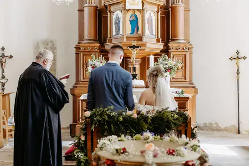 Braut und Bräutigam sitzen Seite an Seite am Altar in einer geschmückten Kirche, einem Priester gegenüber, der während der Hochzeitszeremonie aus einem Buch vorliest. Der Altar ist mit Blumen und religiösen Ikonen geschmückt.