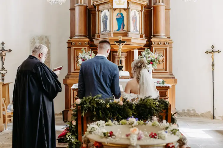 Braut und Bräutigam sitzen Seite an Seite am Altar in einer geschmückten Kirche, einem Priester gegenüber, der während der Hochzeitszeremonie aus einem Buch vorliest. Der Altar ist mit Blumen und religiösen Ikonen geschmückt.