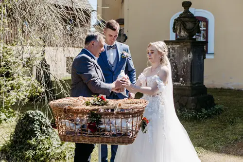 Eine Braut und ein Bräutigam stehen im Freien neben einem Mann mit einem großen Weidenkorb voller Geschenke oder Leckereien und nehmen bei hellem Tageslicht an einer Hochzeitstradition teil.