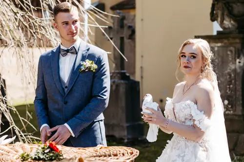 Braut und Bräutigam stehen im Freien in Hochzeitskleidung und halten jeweils eine weiße Taube in den Händen. Vor ihnen steht ein großer Weidenkorb mit weiteren Tauben und Blumen. Sie lächeln und sind von Grün und historischen Gebäuden umgeben.