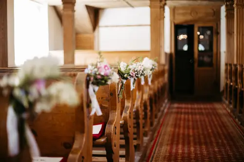 Ein hölzerner Kircheninnenraum mit mit kleinen Blumensträußen geschmückten Kirchenbankreihen, einem rot gemusterten Gangläufer und einer Orgel über dem Eingang im hinteren Bereich.