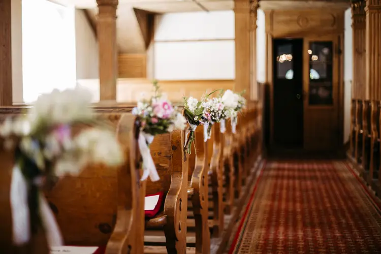 Ein hölzerner Kircheninnenraum mit mit kleinen Blumensträußen geschmückten Kirchenbankreihen, einem rot gemusterten Gangläufer und einer Orgel über dem Eingang im hinteren Bereich.