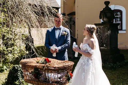 Braut und Bräutigam stehen im Freien in Hochzeitskleidung und halten jeweils eine weiße Taube in den Händen. Vor ihnen steht ein großer Weidenkorb mit weiteren Tauben und Blumen. Sie lächeln und sind von Grün und historischen Gebäuden umgeben.