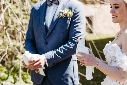 Braut und Bräutigam stehen im Freien und halten jeweils eine weiße Taube in den Händen. Der Bräutigam trägt einen blauen Anzug mit Ansteckblume, die Braut ein weißes Kleid mit Blumenmuster. Beide lächeln im Sonnenlicht.