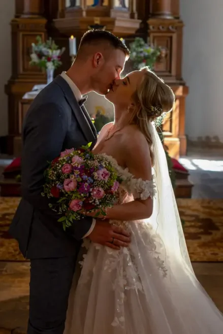 Braut und Bräutigam küssen sich in einer Kirche und stehen eng beieinander. Die Braut trägt ein weißes Kleid und hält einen Strauß rosa und lila Blumen in den Händen. Das Sonnenlicht erhellt das Paar sanft.
