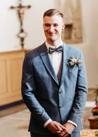 Ein lächelnder Mann in einem blauen Anzug mit Fliege und Blumenanstecker steht im Innenbereich, möglicherweise bei einer Hochzeitszeremonie, mit verschwommenem Kirchendekor im Hintergrund.