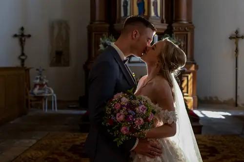 Braut und Bräutigam küssen sich in einer Kirche und stehen eng beieinander. Die Braut trägt ein weißes Kleid und hält einen Strauß rosa und lila Blumen in den Händen. Das Sonnenlicht erhellt das Paar sanft.