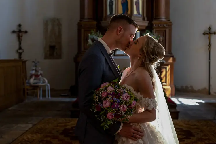 Braut und Bräutigam küssen sich in einer Kirche und stehen eng beieinander. Die Braut trägt ein weißes Kleid und hält einen Strauß rosa und lila Blumen in den Händen. Das Sonnenlicht erhellt das Paar sanft.