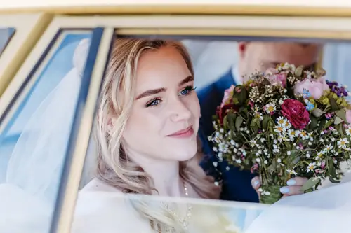 Eine Braut mit blondem Haar sitzt in einem Auto, lächelt sanft und hält einen bunten Blumenstrauß in der Hand, hinter ihr ist eine verschwommene Gestalt zu sehen.
