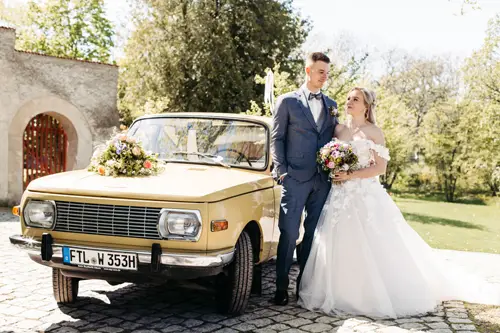 Eine Braut in einem weißen Brautkleid und ein Bräutigam in einem blauen Anzug stehen an einem sonnigen Tag im Freien neben einem gelben, mit Blumen geschmückten Wartburg mit Bäumen und Steingebäuden im Hintergrund.