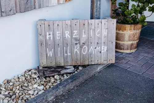 Ein Holzschild mit der handgeschriebenen deutschen Aufschrift „Herzlich Willkommen“ steht auf einem Steinbeet neben einer Topfpflanze und einem gepflasterten Gehweg.