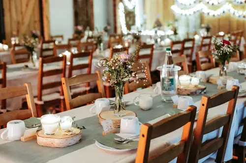 Ein langer, für eine Veranstaltung gedeckter Tisch mit Holzstühlen, Blumengestecken, Kerzen, Tassen, Tellern und Wasserflaschen in einem warm beleuchteten, mit Lichterketten dekorierten Raum.