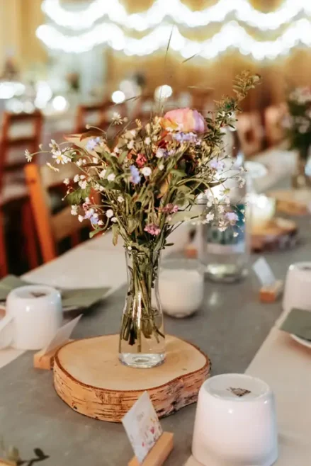 Eine Glasvase mit einem Strauß Wildblumen steht auf einem Tafelaufsatz aus Holz auf einem dekorierten Tisch, umgeben von Tassen, Tischkarten und sanften Lichterketten im Hintergrund.