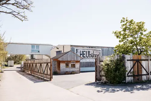 Ein Holztor und ein Zaun öffnen sich zu einem gepflasterten Bereich mit einer kleinen Holzhütte und einem größeren Metallgebäude dahinter, auf dem ein Schild mit der Aufschrift „HEUbühne“ steht. Im Hintergrund sind Bäume und ein klarer Himmel zu sehen.