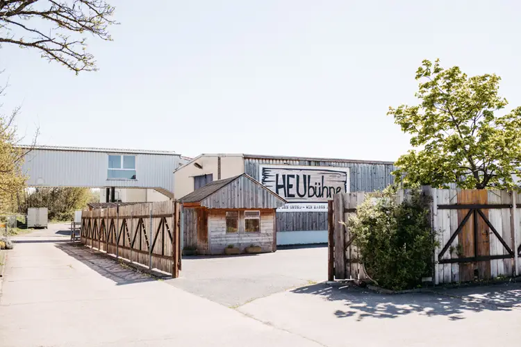 Ein Holztor und ein Zaun öffnen sich zu einem gepflasterten Bereich mit einer kleinen Holzhütte und einem größeren Metallgebäude dahinter, auf dem ein Schild mit der Aufschrift „HEUbühne“ steht. Im Hintergrund sind Bäume und ein klarer Himmel zu sehen.