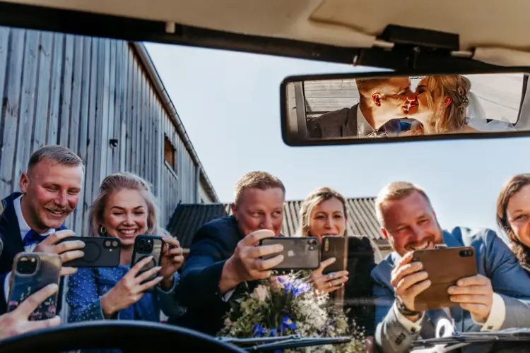 Eine Gruppe lächelnder Menschen fotografiert mit ihren Handys durch ein Autofenster. Im Rückspiegel ist ein küssendes Paar zu sehen, das einen freudigen Hochzeitsmoment festhält.